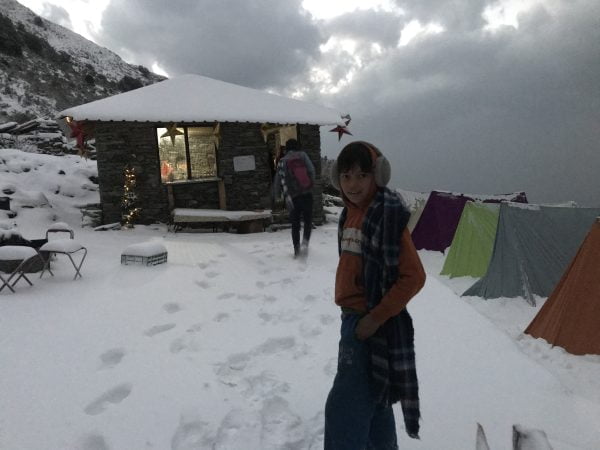 Winter trekking campsite in the Himalayas with snow-covered tents and a trekker wearing earmuffs near a mountain hut Triund trek